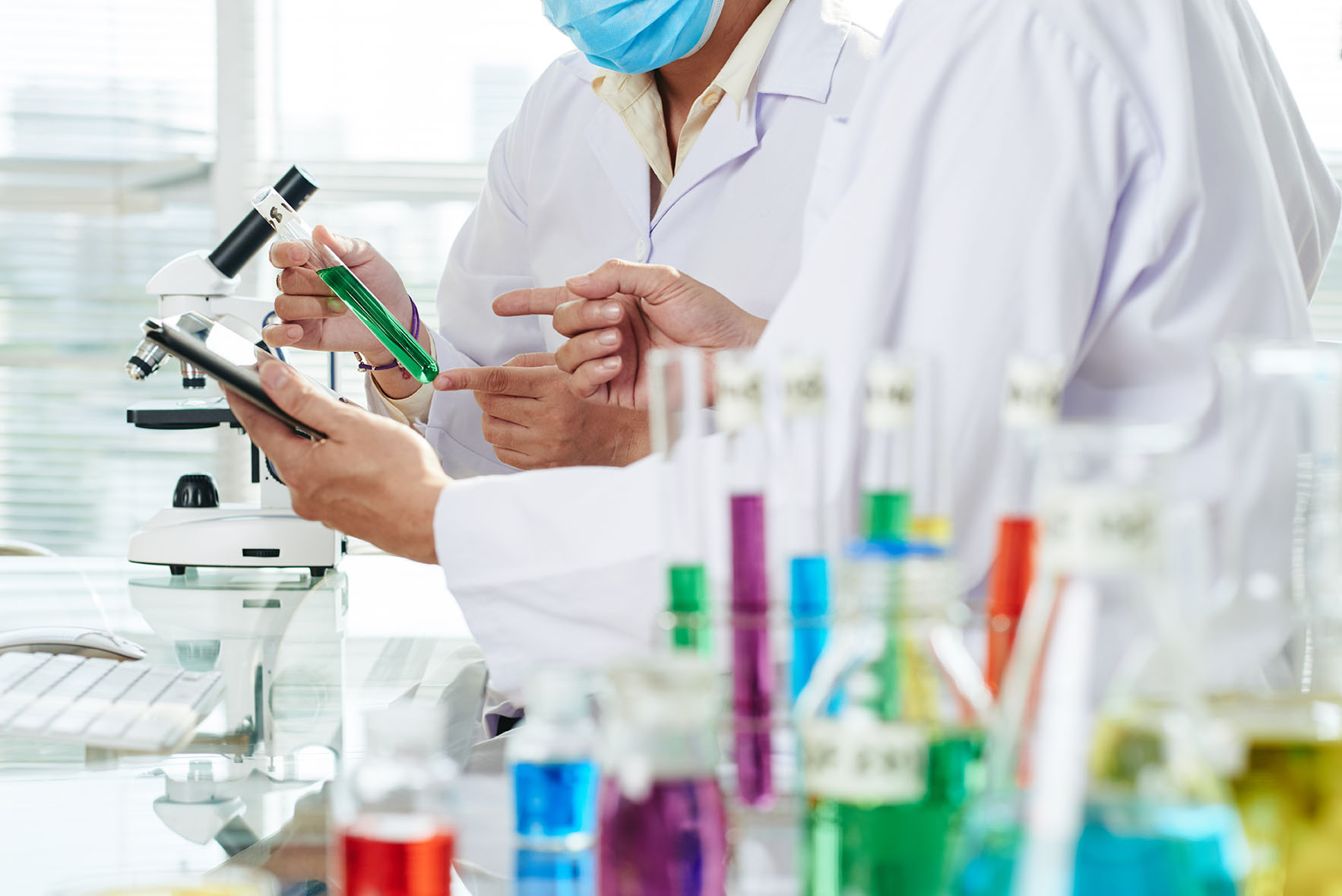 Close-up shot of unrecognizable chemists wearing white coats examining content of test tube and taking necessary notes with help of digital tablet while carrying out experiment